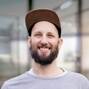 Smiling man with a beard wearing a brown cap and gray shirt, standing in a modern indoor setting.