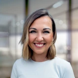 Smiling woman with shoulder-length hair, wearing a light-colored top, in a modern office setting with blurred background.