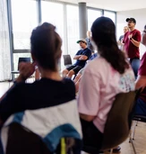 A group of people seated in a circle, listening to a speaker in a casual meeting or workshop setting.