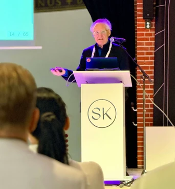 A speaker presents at a podium with a laptop, addressing an audience in a conference setting. A presentation slide is visible in the background.